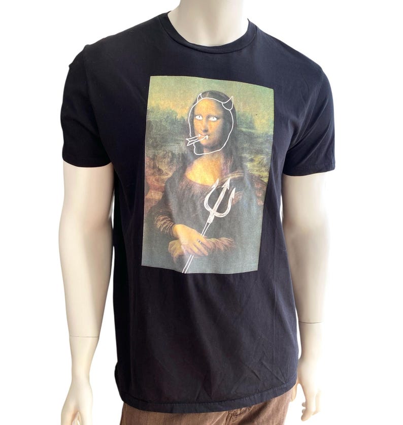 May include: Black t-shirt featuring a humorous graphic of the Mona Lisa with devil horns, a cigarette, and a trident. The graphic is a parody of the famous painting. The shirt is made of a soft material.