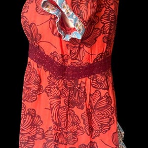May include: A red top with a floral pattern in dark brown. The top features a burgundy lace detail at the waist and blue and white patterned trim on the sleeves and hem. The short-sleeved blouse is a women's apparel item.