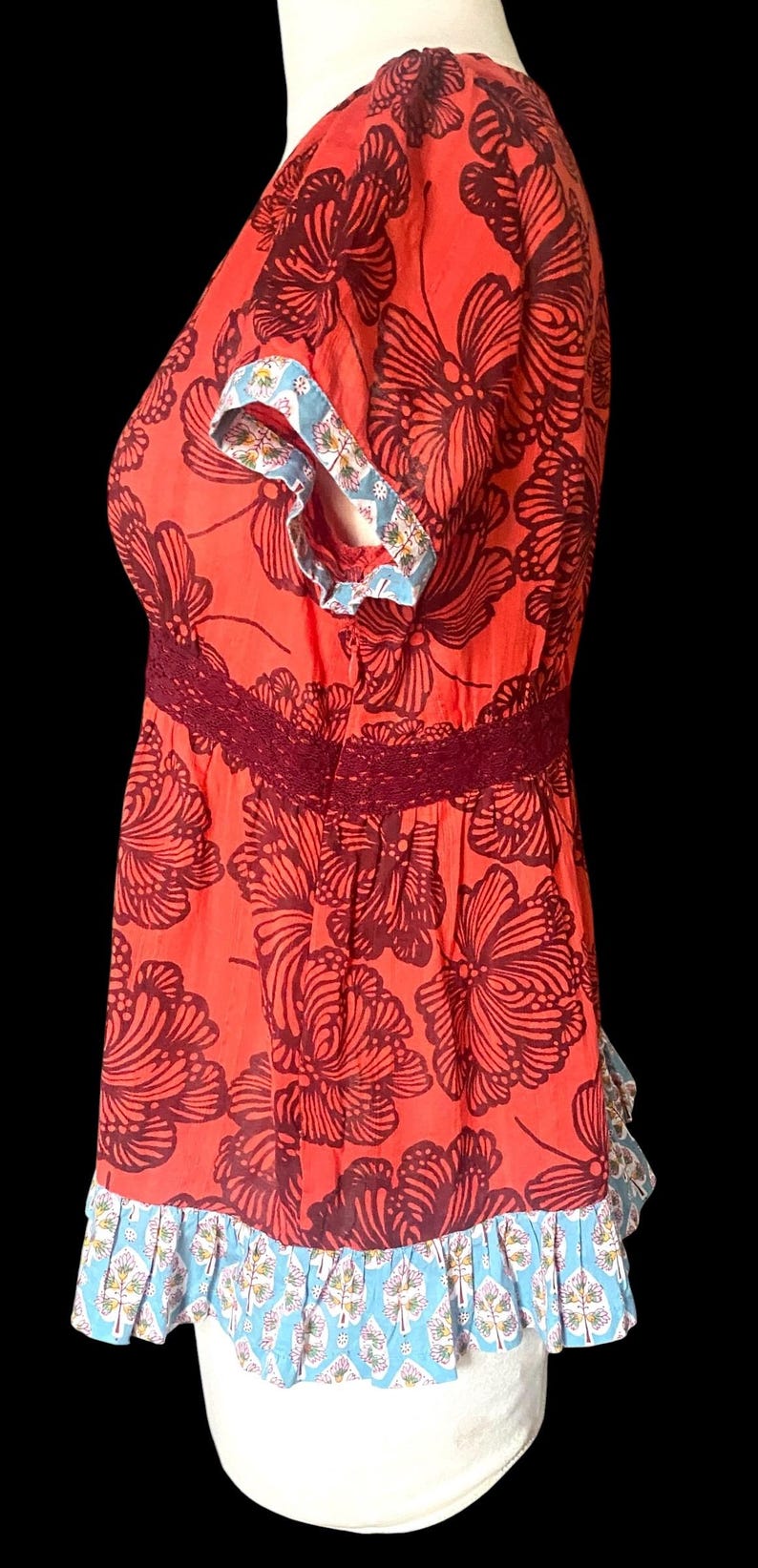 May include: A red top with a floral pattern in dark brown. The top features a burgundy lace detail at the waist and blue and white patterned trim on the sleeves and hem. The short-sleeved blouse is a women's apparel item.