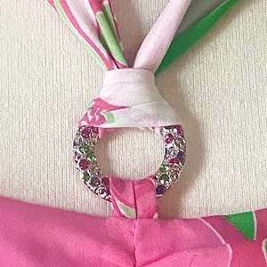 Op de afbeelding: Close-up van een roze haltertop met een decoratieve zilveren ring. De ring is bezet met kleurrijke strass-steentjes. De halterbandjes zijn bedrukt met roze, witte en groene bloemmotieven.