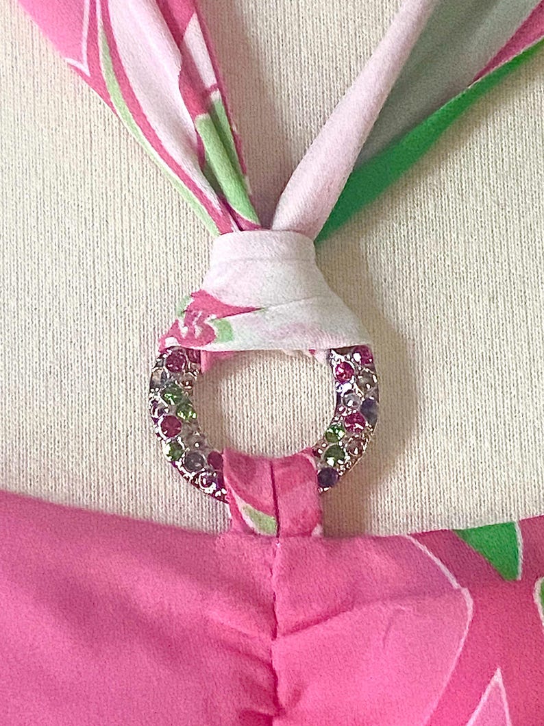 Op de afbeelding: Close-up van een roze haltertop met een decoratieve zilveren ring. De ring is bezet met kleurrijke strass-steentjes. De halterbandjes zijn bedrukt met roze, witte en groene bloemmotieven.