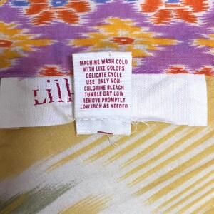 May include: Close-up of a colorful fabric with a tag. The fabric features a pattern of orange, purple, and yellow. The tag reads "Lill" and includes washing instructions: machine wash cold, delicate cycle, tumble dry low.