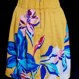 Op de afbeelding: Een gele rok met een bloemenpatroon. De rok heeft een textuurstof en een ritssluiting. Het bloemenpatroon bevat blauwe, roze en witte bloemen.