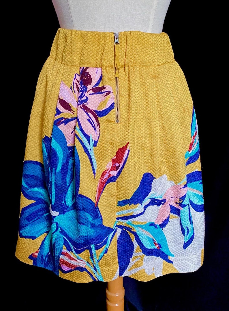 Op de afbeelding: Een gele rok met een bloemenpatroon. De rok heeft een textuurstof en een ritssluiting. Het bloemenpatroon bevat blauwe, roze en witte bloemen.