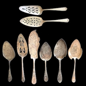 May include: A collection of eight silver-toned metal cake servers and pastry knives arranged in two rows. Each utensil features unique decorative engravings, intricate cut-out patterns, or embossed floral motifs on the blades and handles.
