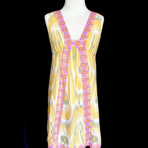 May include: A sleeveless, knee-length dress with a V-neck. The dress features a yellow, white, and blue ikat pattern. The neckline and vertical seams are trimmed with a pink border and orange floral accents. The hemline also has a pink border.
