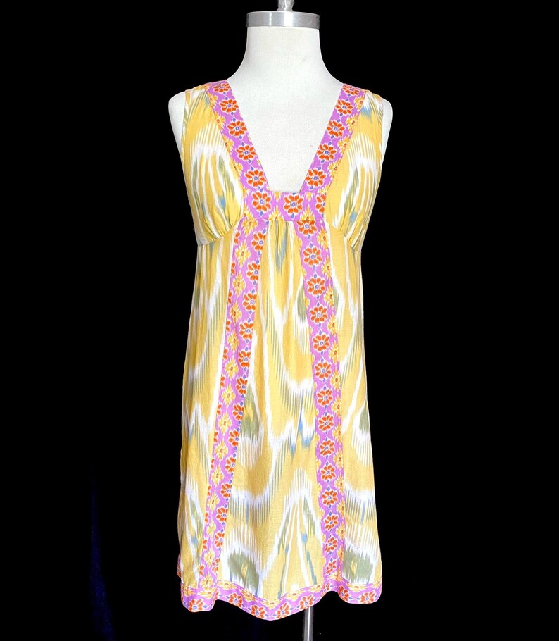 May include: A sleeveless, knee-length dress with a V-neck. The dress features a yellow, white, and blue ikat pattern. The neckline and vertical seams are trimmed with a pink border and orange floral accents. The hemline also has a pink border.
