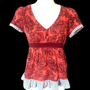 May include: Coral-colored top with a V-neck and short sleeves. The top features a large floral pattern in a darker shade of red. A dark red lace detail accents the waist, and the sleeves and hem are trimmed with a blue and white patterned fabric.