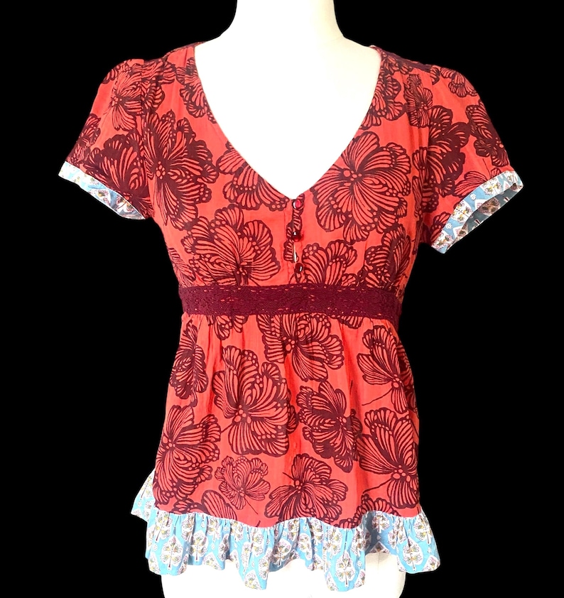 May include: Coral-colored top with a V-neck and short sleeves. The top features a large floral pattern in a darker shade of red. A dark red lace detail accents the waist, and the sleeves and hem are trimmed with a blue and white patterned fabric.
