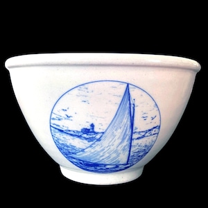 May include: White ceramic bowl with a blue sailboat and lighthouse design. The nautical scene is inside a blue circle. The bowl has a wide rim and a slightly flared shape, perfect for serving.