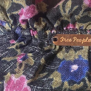 May include: Close-up of a floral fabric with pink, blue, and gold flowers on a dark background. A small, brown metal tag with the words "Free People" is attached to the fabric.