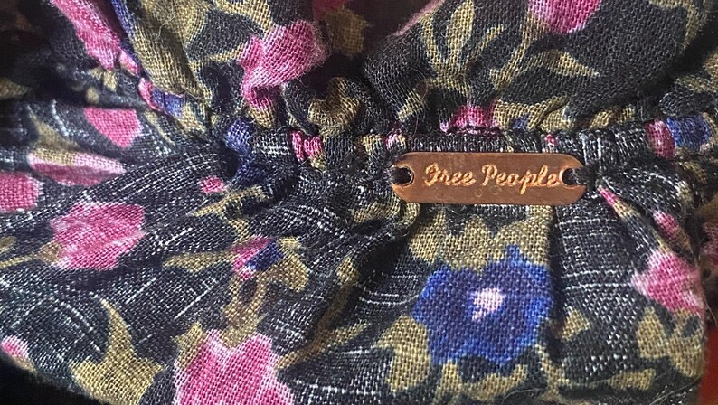 May include: Close-up of a floral fabric with pink, blue, and gold flowers on a dark background. A small, brown metal tag with the words "Free People" is attached to the fabric.