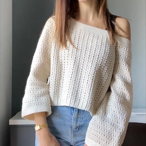 May include: Off-the-shoulder cream-colored crochet sweater with long sleeves. The sweater is paired with light blue jeans. The crochet pattern is visible, and the sleeves have a flared design. The sweater is a casual, handmade garment.