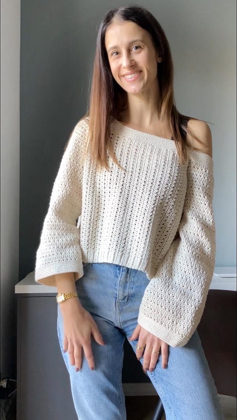 May include: Off-the-shoulder cream-colored crochet sweater with long sleeves. The sweater is paired with light blue jeans. The crochet pattern is visible, and the sleeves have a flared design. The sweater is a casual, handmade garment.