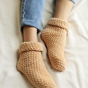 Pu&ograve; includere: Un paio di calzini da pantofola in maglia beige morbidi con una trama a coste. I calzini sono indossati da una persona seduta su una coperta bianca.