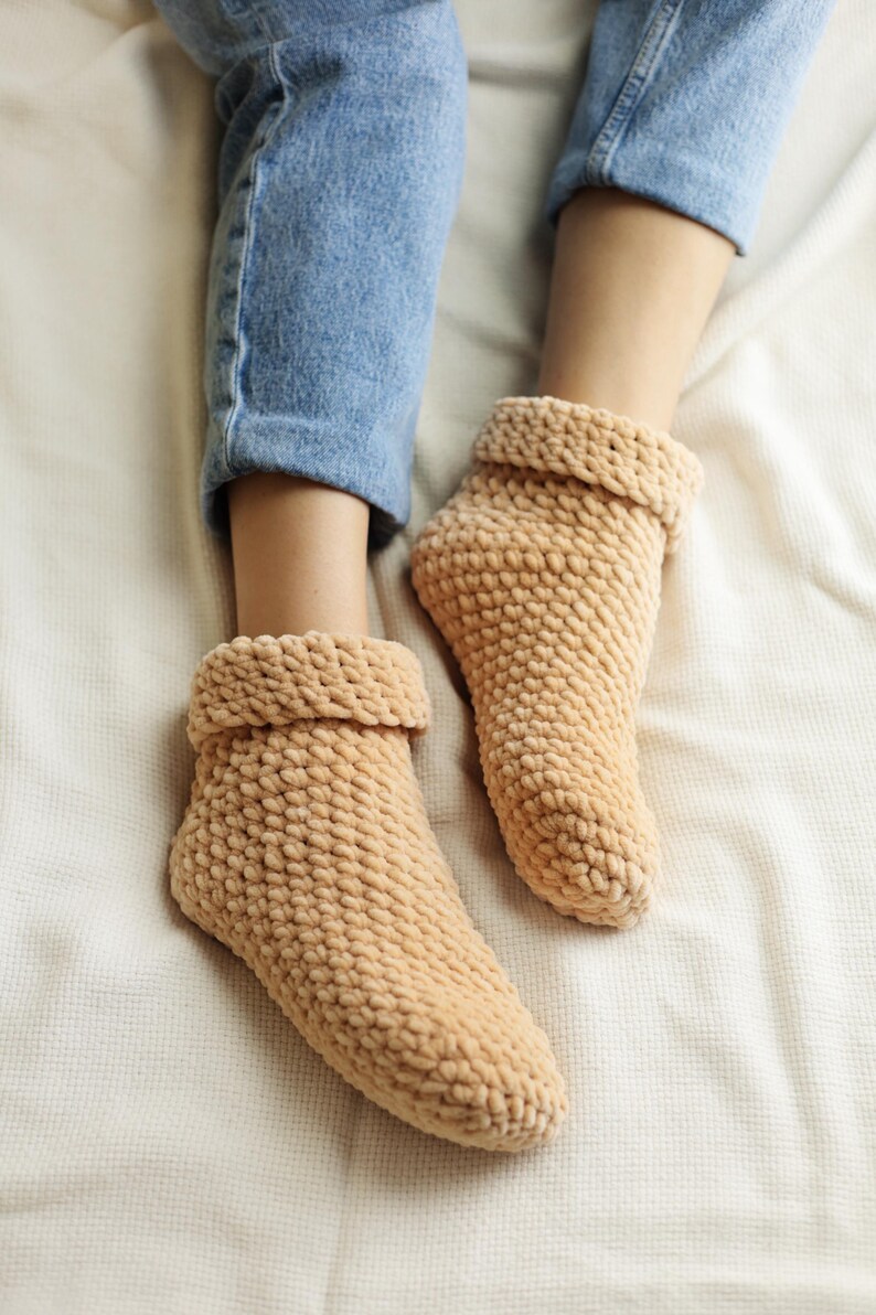 Pu&ograve; includere: Un paio di calzini da pantofola in maglia beige morbidi con una trama a coste. I calzini sono indossati da una persona seduta su una coperta bianca.