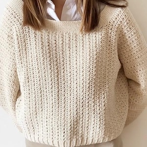 May include: Cream-colored, hand-crocheted sweater with a textured stitch pattern. The sweater has a wide neckline and long sleeves. The model is wearing a white collared shirt underneath and tan pants. The sweater is a cozy, handmade garment.