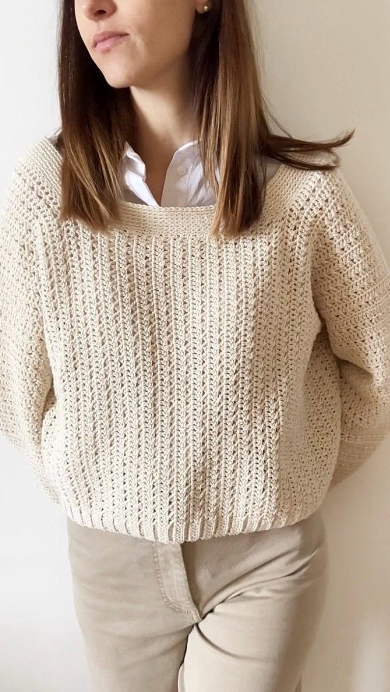 May include: Cream-colored, hand-crocheted sweater with a textured stitch pattern. The sweater has a wide neckline and long sleeves. The model is wearing a white collared shirt underneath and tan pants. The sweater is a cozy, handmade garment.