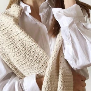 May include: A cream-colored, crocheted shoulder bag with a textured pattern. The bag is draped over a white button-down shirt. The crochet work appears to be made of a soft, natural fiber, and the bag's design is simple and elegant.
