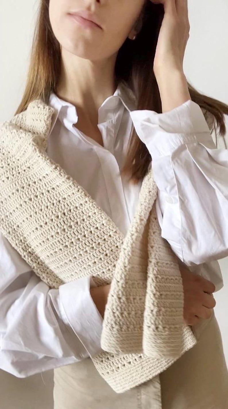 May include: A cream-colored, crocheted shoulder bag with a textured pattern. The bag is draped over a white button-down shirt. The crochet work appears to be made of a soft, natural fiber, and the bag's design is simple and elegant.