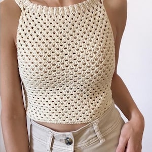 May include: Cream-colored, handmade crochet top with a square neckline and open weave design. The top is paired with beige pants. The crochet top is a sleeveless garment, perfect for warm weather. The overall style is casual and bohemian.