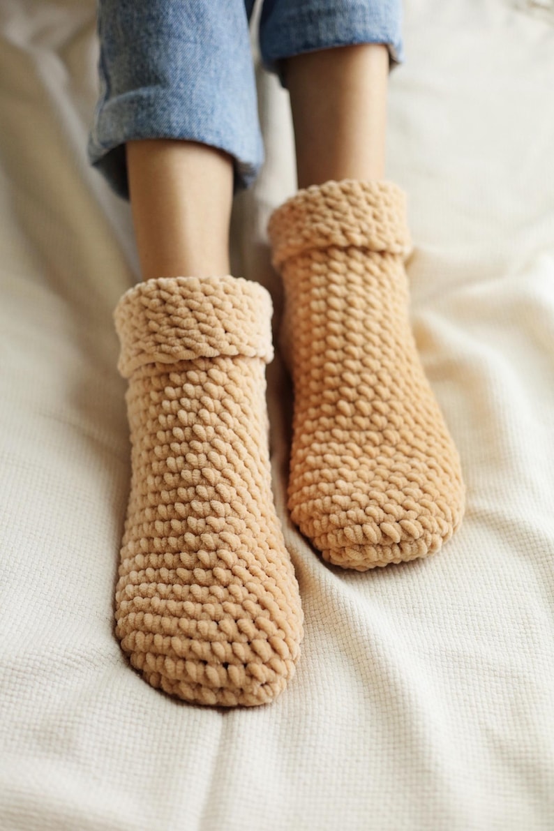 Pu&ograve; includere: Un paio di calzini da pantofola in maglia marrone e confortevoli con una superficie testurizzata. I calzini sono indossati da una persona seduta su una coperta bianca.