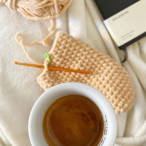 Pu&ograve; includere: Una tazza di ceramica bianca con un espresso marrone al suo interno. La tazza ha le parole "Vintage Collection" stampate sul lato. La tazza &egrave; tenuta da una mano su una coperta bianca con un progetto all'uncinetto e un taccuino nero.