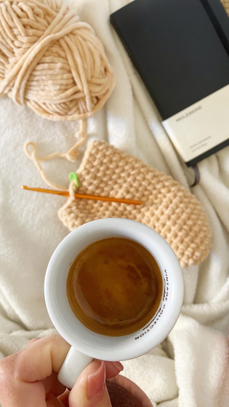 Pu&ograve; includere: Una tazza di ceramica bianca con un espresso marrone al suo interno. La tazza ha le parole "Vintage Collection" stampate sul lato. La tazza &egrave; tenuta da una mano su una coperta bianca con un progetto all'uncinetto e un taccuino nero.