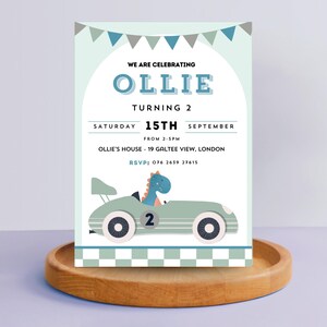 K&ouml;nnte beinhalten: Eine blaue und gr&uuml;ne Geburtstagseinladung mit einem Comic-Dinosaurier, der ein Rennauto f&auml;hrt. Die Einladung lautet "We are celebrating Ollie turning 2" und enth&auml;lt Datum, Uhrzeit und Ort der Party.
