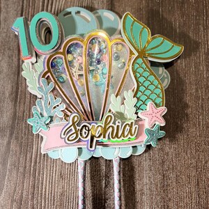 Op de afbeelding: Een cake topper met zeemeermint thema met het nummer 10 in teal. De topper heeft een zeeschelp met een shaker element, een zeemeerminstaart en de naam "Sophia" op een roze banner. Decoratieve elementen zijn koraal, zeesterren en bellen.