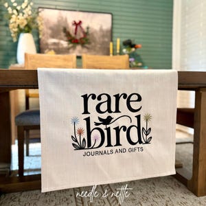 May include: Cream-colored table runner with the words "rare bird" in black script, a bird graphic, and the text "JOURNALS AND GIFTS." The runner has a floral design with colorful accents and the words "needle & nettle" in script.