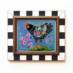 Peut inclure: Une peinture encadr&eacute;e d'une poule noire avec des d&eacute;tails color&eacute;s, debout dans un champ de fleurs roses sur un fond bleu. Le cadre est &agrave; carreaux noir et blanc.