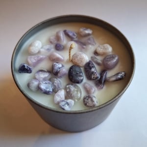 Puede incluir: Una vela en un recipiente de metal gris llena de cera blanca y adornada con varias piedras moradas y grises. Una sola mecha sin encender está centrada en la vela, creando una estética natural y decorativa.