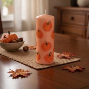 Puede incluir: Una vela de pilar cilíndrica de color melocotón con un patrón repetitivo de calabazas naranjas pintadas a mano con tallos verdes. La vela tiene una mecha blanca centrada en la parte superior y un acabado de cera suave.