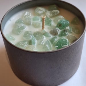 May include: A grey metal container candle with white wax and green gemstones. A single, unlit wick is in the centre. The gemstones are various shades of green and are scattered throughout the wax.