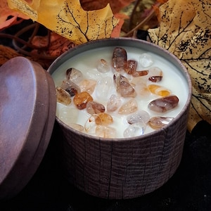 May include: A round, brown candle in a wooden-textured container, topped with clear and amber-coloured crystals. The lid is open, revealing the white wax. Autumn leaves in shades of yellow and orange are in the background.