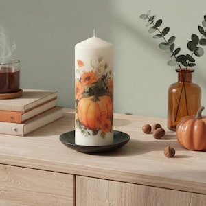 May include: A cylindrical off-white pillar candle featuring a printed design of a large orange pumpkin surrounded by orange and yellow flowers and green foliage, resting on a small black circular plate.
