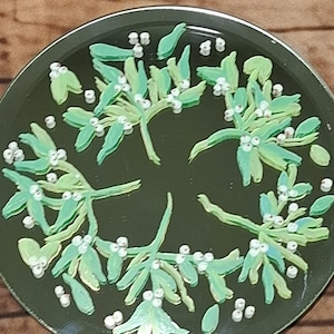 Pode incluir: Um prato decorativo redondo, verde escuro, com um desenho de visco. O visco é pintado em tons de verde e branco, com pequenas bagas brancas. O prato tem uma borda metálica e está sobre um fundo de madeira.