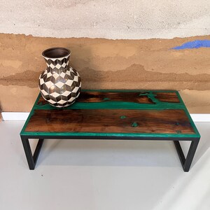 May include: A dark wood coffee table with a green epoxy river design and a black metal frame. A black and white geometric vase sits on the table.