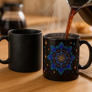 May include: Two black ceramic mugs, one with a mandala design in blue, teal, and gold, the other plain. Coffee is being poured into the decorated mug. The mugs are on a wooden surface.