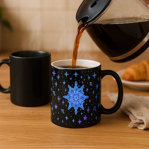 Puede incluir: Una taza de cerámica negra con un diseño celestial se llena de café de una jarra de vidrio. La taza presenta un patrón de estrellas azules y moradas y un diseño floral central. Otra taza negra es visible.