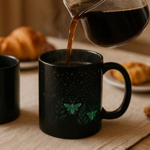 May include: A black ceramic mug featuring a celestial design of constellations and green fireflies. Hot coffee is being poured into the mug from a glass carafe. The mug has a curved handle and is on a beige surface.