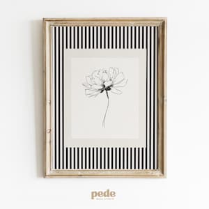 May include: Framed floral art print. The artwork features a delicate black and white flower illustration on a light background, set within a striped border of black and white. The frame is a light gold color.