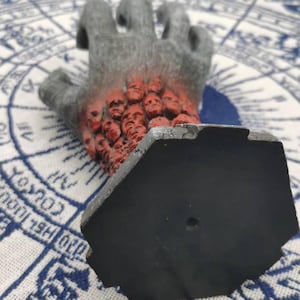 May include: A black hand sculpture with a hexagonal base. The hand is made of a dark gray material and has a red, fleshy texture. The hand is covered in small, red faces.