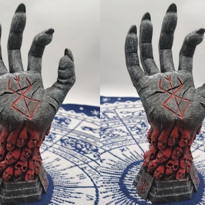 May include: A gray hand sculpture with a red symbol on the palm. The hand is holding a base filled with red faces.