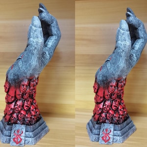 May include: A gray hand sculpture with a textured surface. The hand is holding a collection of small, red faces. The sculpture is on a gray base with a red symbol.