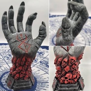 May include: A gray hand sculpture with a red symbol on the palm. The hand is made up of many small faces and skulls. The sculpture is on a black base.