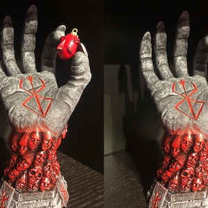 May include: A gray hand sculpture with a red symbol on the palm, holding a red Christmas ornament. The hand is made up of many small faces, all with red eyes and mouths.