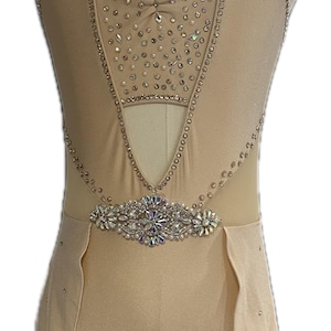 Könnte beinhalten: Ein hautfarbener Tanz-Leotard mit einem transparenten Einsatz und einem Gürtel mit Schmucksteinen. Der Leotard hat einen Halterneck und einen hohen Beinausschnitt. Der Rock ist am Leotard befestigt und hat einen gewellten Saum.