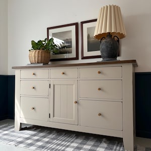 May include: A cream-colored wooden dresser with multiple drawers and a central cabinet. A brown countertop supports a lamp with a beige shade, two framed pictures, and a wicker basket with a green plant. The dresser has gold-colored knobs.